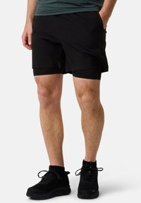 Zwarte sportshorts van een gladde buitenstof, voorzien van een aansluitende binnenvoering, zijzakken en een lengte tot halverwege de dijen. Gecombineerd met zwarte sneakers.