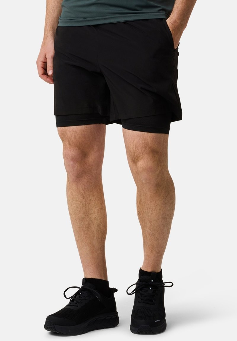 Zwarte sportshorts van een gladde buitenstof, voorzien van een aansluitende binnenvoering, zijzakken en een lengte tot halverwege de dijen. Gecombineerd met zwarte sneakers.
