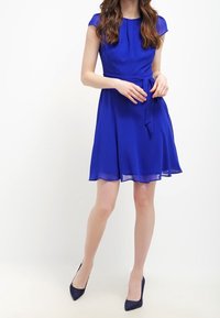 Kort blå klänning med figurnära liv, svängande kjol, korta ärmar och ett knytbälte i midjan, gjord av ett mjukt och lätt material.