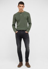 Mężczyzna w oliwkowej, długiej koszuli, ciemnych dżinsach i brązowych butach na sznurówki, stojący z rękami w kieszeniach na białym tle.