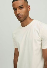 Kortärmad T-shirt med rundad hals i ljusbeige, tillverkad av mjukt bomullstyg med en avslappnad passform och minimala sömmar för ett rent utseende.