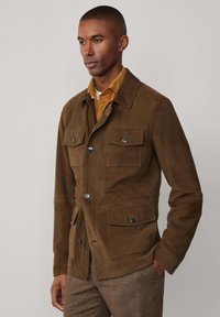 Braune Wildlederjacke mit Kragen, ausgestattet mit zwei Brusttaschen und zwei Seitentaschen, Knopfverschluss und einer glatten Textur.