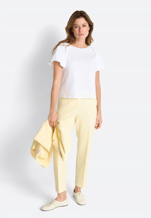 Žena s vlnitými vlasmi, oblečená v bielom tričku s krátkym rukávom, svetložltých nohaviciach a bielych topánkach, držiaca žltú bundu v rovnakej farbe na bielom pozadí.