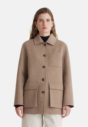 Mujer con abrigo de lana beige abotonado, con tres grandes bolsillos y cuello, combinado con pantalones claros y pendientes de aro.
