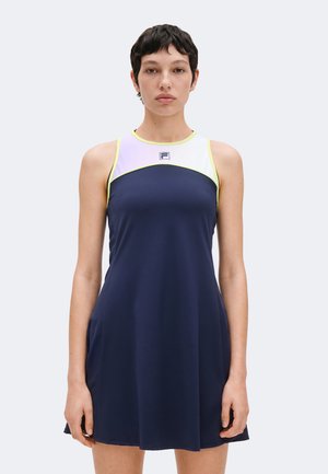 Jeune femme aux cheveux courts et foncés, portant une robe sans manches bleu marine et blanche avec une bordure jaune, debout face à l'avant sur un fond uni.