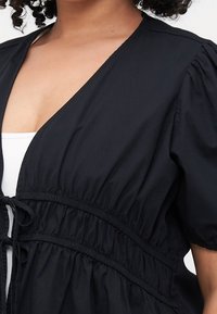 Schwarze, leichte Bluse mit tiefem V-Ausschnitt, Puffärmeln und gerafftem Bund mit Kordelzug. Glatte Textur und minimalistische Designdetails.