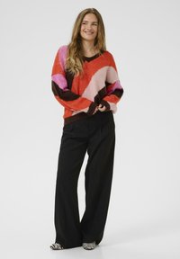 Pull coloré avec un motif abstrait dans les tons orange, rose et marron, avec des poignets marron, associé à un pantalon noir ample et des chaussures rayées.