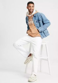 Dżinsowa kurtka, beżowy hoodie z tekstem, białe spodnie i jasne sneakersy. Model pozuje na stołku, prezentując swobodny styl uliczny.