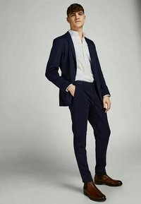 Jovem homem de pé, confiante, vestido com um fato azul-marinho, camisa branca e botas de couro castanho, contra um fundo liso cinza-claro.