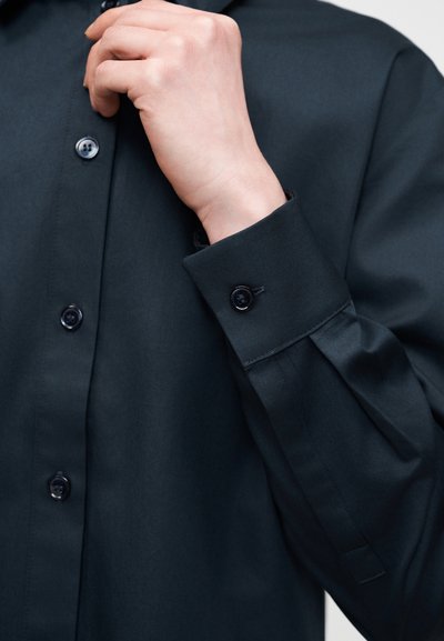 Χέρι που ρυθμίζει τον γιακά μαύρου πουκάμισου με μακριά μανίκια, κουμπιά και λεπτομέρεια μανσετών ορατά.