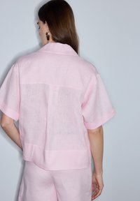 Mujer con cabello largo y castaño que lleva una camiseta rosa claro transparente de manga corta y pantalones a juego, de pie de espaldas a la cámara sobre un fondo liso.