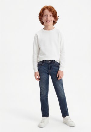 Garçon roux souriant portant un sweat-shirt blanc, un jean bleu et des baskets blanches, debout sur un fond blanc.