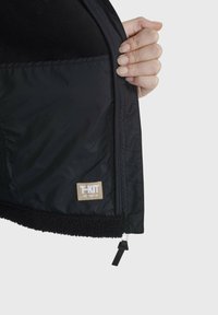 Unutrašnjost crne jakne s glatkim materijalom i pahuljastom podstavom, s vidljivom oznakom "T-KIT" blizu zatvarača.