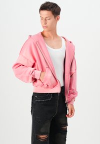 Lyserød cropped hoodie med lynlås fortil, oversized pasform og ribbede manchetter, båret over en hvid tank top, parret med slidte sorte jeans.