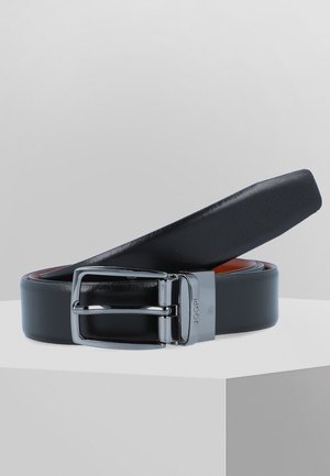 Schwarzer Ledergürtel mit silberner Metallschnalle, aufgerollt und auf einer weißen Oberfläche vor einem schlichten grauen Hintergrund liegend.