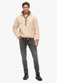 Polarowy sweter, w kolorze kremowym, z pół-zipem, kieszeń na piersi z zamkiem, dwie kieszenie z przodu, nałożony na czerwoną koszulę w kratę, zestawiony z szarymi dżinsami.