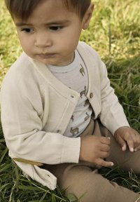 Helle beigefarbene Strickjacke mit Knopfverschluss, über einem grauen Shirt mit kleinen Tiermotiven und braunen Ripp-Hosen. Grüne Wiese im Hintergrund.