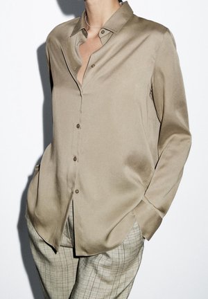 Chemise en satin beige à boutons, associée à un pantalon à carreaux gris, mains dans les poches, sur un fond blanc uni.