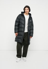 Abrigo acolchado negro con capucha y cremallera, con paneles acolchados, un pequeño logo en el pecho, combinado con pantalones verde oliva y zapatillas blancas.