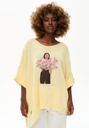 Kobieta z kręconymi włosami nosi luźny żółty sweter z ilustracją kobiety z różowymi kwiatami na torsie i białe spodnie.