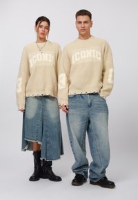 Beige knit sweaters with "ICONIC" and "3" printed in white, paired with faded denim skirts and jeans, and black ankle boots. Textured fabric details.