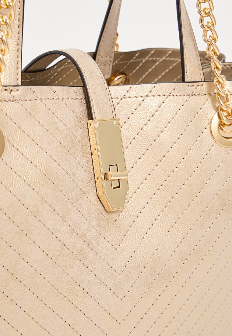 Bolso tote de cuero dorado con un patrón en chevrón, que presenta un cierre dorado y asas de cadena. Textura suave con sutiles detalles de costura.