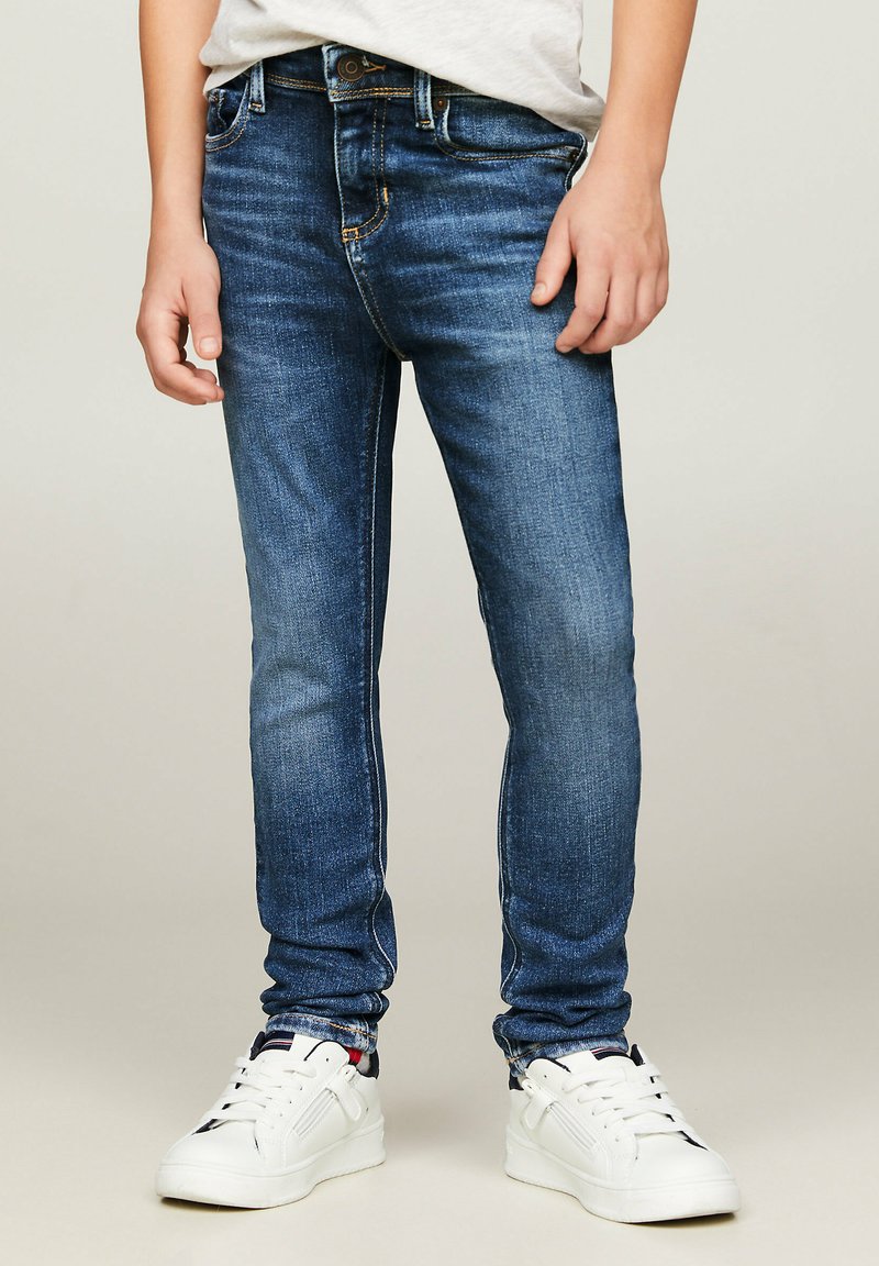 Wąskie niebieskie dżinsy z lekko wyblakłym wykończeniem, z pięcioma kieszeniami i kontrastowym przeszyciem, zestawione z białymi trampkami.