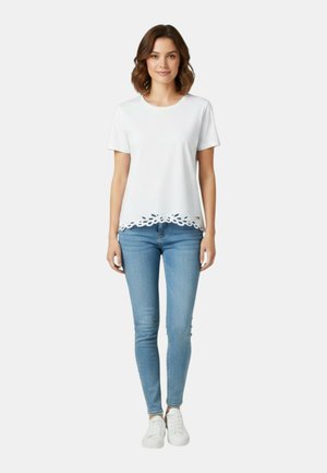 Donna in piedi che indossa una maglietta bianca a maniche corte con orlo traforato a fiori, jeans skinny azzurri e scarpe da ginnastica bianche su sfondo semplice.