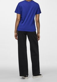 Persona in piedi di spalle, con una maglietta blu a maniche corte, pantaloni neri a gamba larga e sneakers nere con suole bianche.