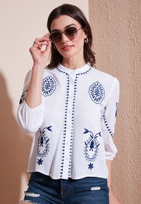 Blouse blanche avec broderies bleues, col rond, bouton devant, manches trois-quarts et motifs floraux. Le modèle porte des lunettes de soleil.