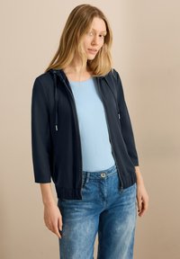Giacca zip-up blu navy con cappuccio, maniche a tre quarti e orlo a coste. Indossata sopra una maglietta azzurro chiaro, abbinata a jeans in denim chiaro.