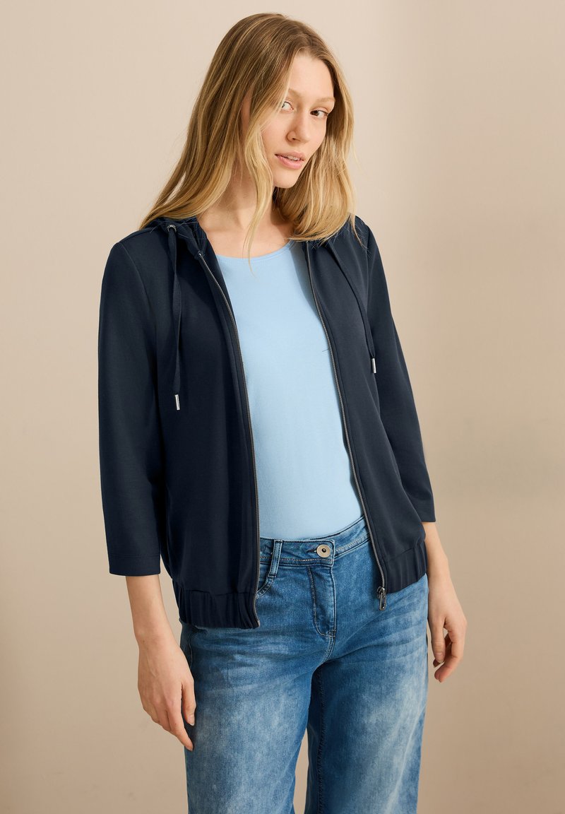 Giacca zip-up blu navy con cappuccio, maniche a tre quarti e orlo a coste. Indossata sopra una maglietta azzurro chiaro, abbinata a jeans in denim chiaro.