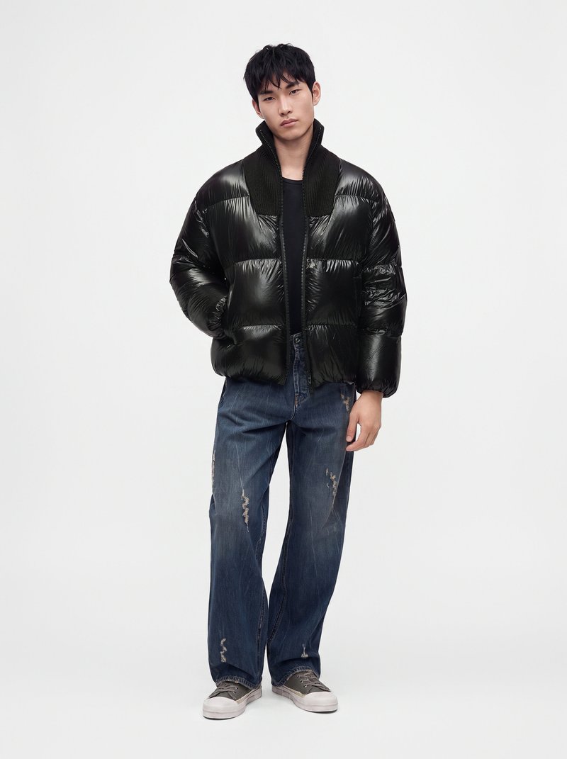 Jeune homme avec une doudoune noire, une chemise noire, un jean bleu délavé ample et des baskets grises sur un fond clair uni.