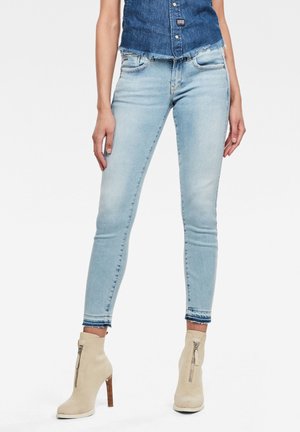 Nainen, joka pukeutunut vaaleansinisiin kapealahkeisiin farkkuihin, joissa on kärjestä repsottavat reunat, beigeihin korkeakorkoisiin nilkkureihin, joissa on vetoketjut edessä, ja sleeveless farkkutoppiin.