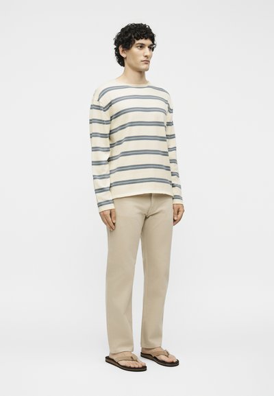 Homme debout portant une chemise crème à manches longues avec des rayures grises, un pantalon beige et des sandales beiges sur un fond blanc uni.