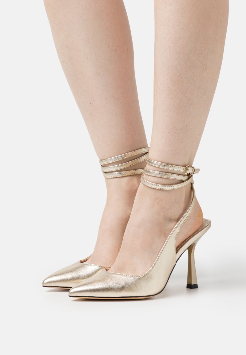 Chaussures à talons hauts métalliques dorées avec un bout pointu et des brides à la cheville. Présentent un design élégant et une finition texturée.
