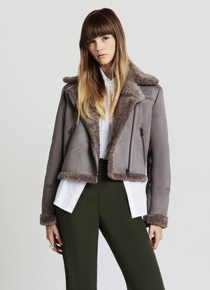 Giovane donna con lunghi capelli castano chiaro indossa una giacca shearling grigia sopra una camicia bianca e pantaloni verde scuro, in piedi contro uno sfondo neutro.