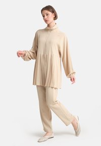 Completo beige a due pezzi con tunica plissettata e pantaloni slim, caratterizzato da un colletto alto e una zip, realizzato in un tessuto morbido e liscio.
