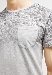 Detailní záběr světle šedého trička s propracovaným černým květinovým a paisley vzorem kolem hrudi, ramene a rukávu, se jednoduchou náprsní kapsou.