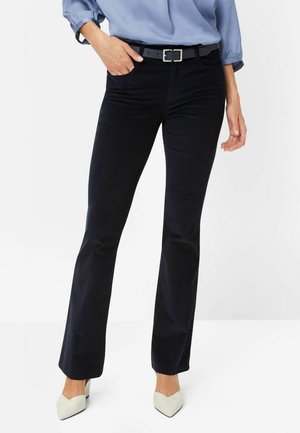 Mujer con pantalones acampanados negros con cinturón negro, blusa azul claro y tacones blancos de punta puntiaguda, de pie contra un fondo blanco.