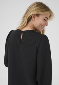 Černá blůza s texturovanou tkaninou, která má otvor na zádech ve tvaru klíče a dlouhé rukávy s nabranými rameny. Hladký, elegantní design.