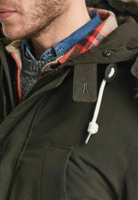 Parka vert olive avec fermeture éclair et boutons, doublure en polaire visible, col en tricot gris et accents en corde blanche sur la capuche.