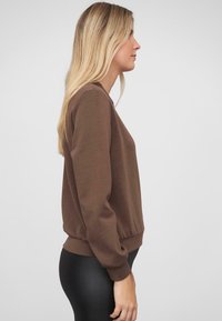 Brauner Pullover mit langen Ärmeln, rundem Ausschnitt, elastischem Bund und glatter Textur. Schlank geschnittenes Design, kombiniert mit schwarzen Leggings.