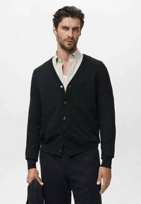 Svart cardigan i mjukt material, med V-hals, långa ärmar och knäppning med knappar. Bärs över en ljus skjorta, ihop med mörka byxor.
