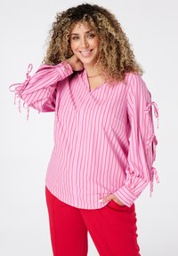 Roze en witte gestreepte blouse met een V-hals en gathered mouwen met linten. Gepaarde met rode broeken. Glad textuur.