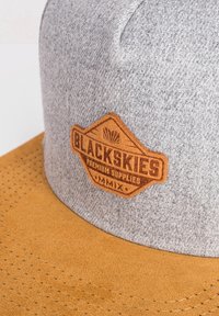 Grå tygmössa med en brun mockaskärm. Har ett broderat emblem med texten "BLACKSKIES PREMIUM SUPPLIES" i hexagonal form.