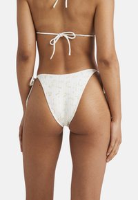 Biele bikiny s ryhovanou štruktúrou a kvetinovým vzorom. Vrchná časť sa viaže vzadu, zatiaľ čo spodná časť má bočné viazanie a vysoký strih.
