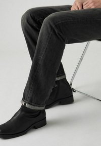 Donkergrijze denim jeans met een lichte was, met omgeslagen broekspijpen die een lichtgrijze onderkant onthullen. G gedragen met zwarte leren enkellaarsjes.