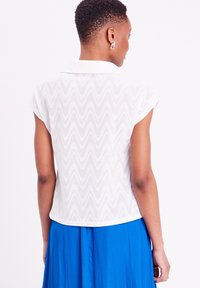 Blouse blanche à manches courtes avec un motif en vagues texturé, à col et à coupe décontractée, portée sur une jupe bleue.