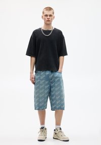 Sort ribbet oversized t-shirt, lyseblå denim shorts med gentagende logo-tryk og beige høj-top sneakers. Kædehalssmykke som accent.